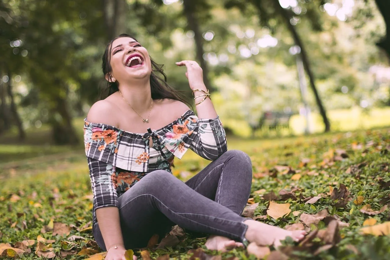 Une jeune femme rieuse assise dans un parc, entourée de feuilles mortes, avec des arbres en arrière-plan.