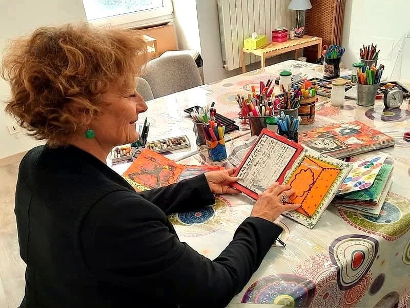 Une femme âgée regarde un livre de souvenirs ou un album photo à une table décorée avec des marqueurs, des fournitures d'art et des livres.