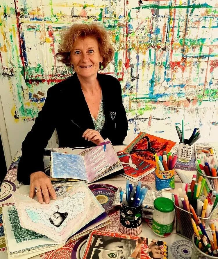 Femme souriante assise à un bureau rempli de fournitures artistiques, avec des œuvres d'art abstraites en arrière-plan.