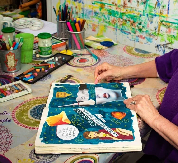 Une personne, portant une manche violette, manipule un magazine ouvert illustré avec des images colorées et du texte. La table autour est remplie de matériel d'art comme des crayons, des marqueurs, et des pots de peinture, dans un espace créatif.