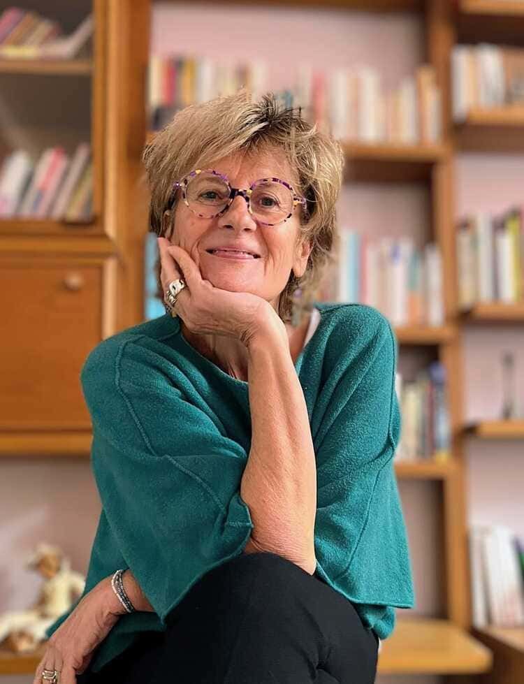 Une femme âgée souriante, portant des lunettes violettes, assise dans une bibliothèque.
