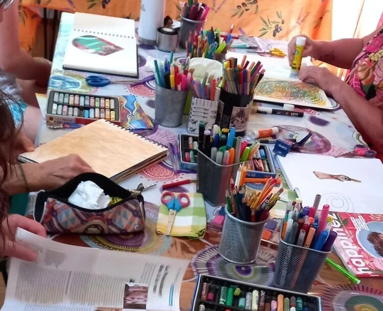 Un tableau de table avec de nombreux crayons, peintures et fournitures artistiques, ainsi que des personnes travaillant ou créant des œuvres.