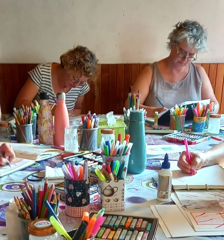 Deux femmes adultes assises à une table remplie de fournitures d'art et de dessin, colorants, feutres, crayons, et papier, en train de dessiner ou de colorier.