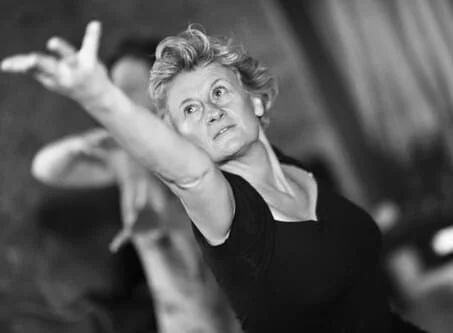 Une femme âgée avec des cheveux courts, faisant un mouvement avec le bras dans une pièce.