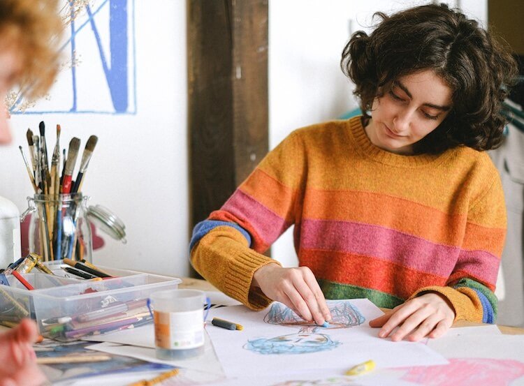 Fille en pull rayé coloré dessinant sur une feuille, avec des pinceaux et des fournitures d'art autour, dans un atelier.