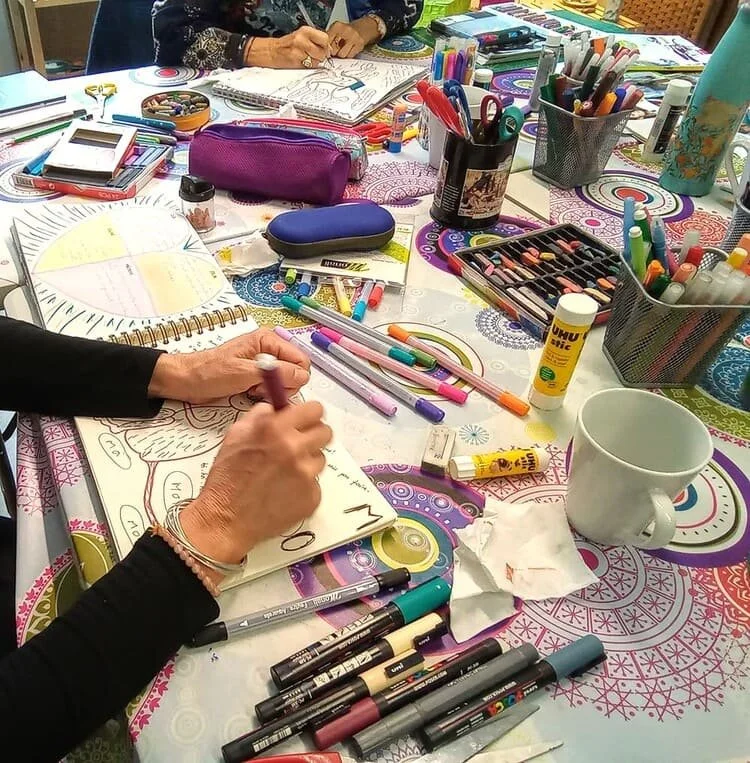 Une table colorée avec des fournitures d'art comme des marqueurs, stylos, gommes, pinceaux, et un carnet de croquis. On voit une personne en train de dessiner ou d'écrire.