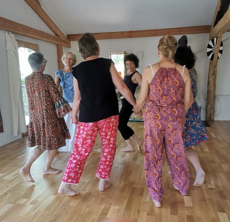 Groupe de sept femmes dansant côte à côte dans une pièce avec un sol en bois et des murs blancs, portant des vêtements colorés et décontractés.