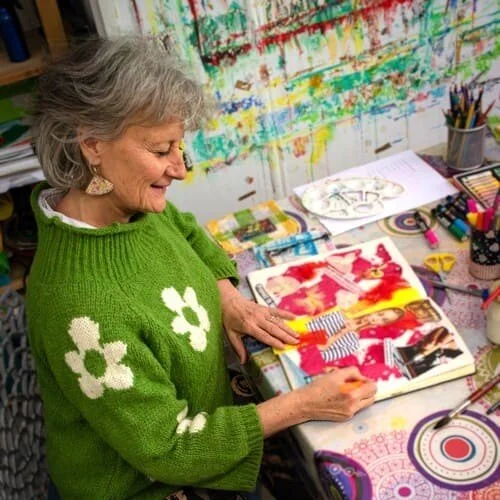 Une femme âgée avec un pull vert à motifs de fleurs blanches, en train de colorier dans un livre d'art à un atelier créatif.