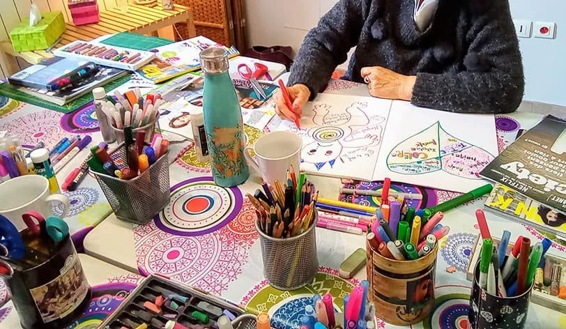 Une personne en train de dessiner sur une table chargée de fournitures artistiques colorées, entourée de marqueurs, crayons, stylos, et autres outils créatifs.