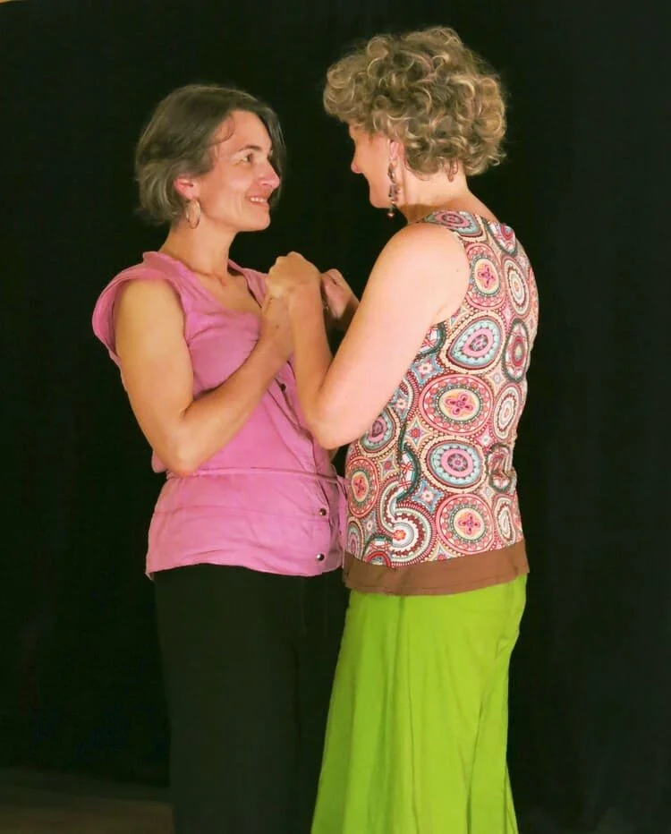 Deux femmes se regardent tendrement, se tenant par la main, contre un fond noir.