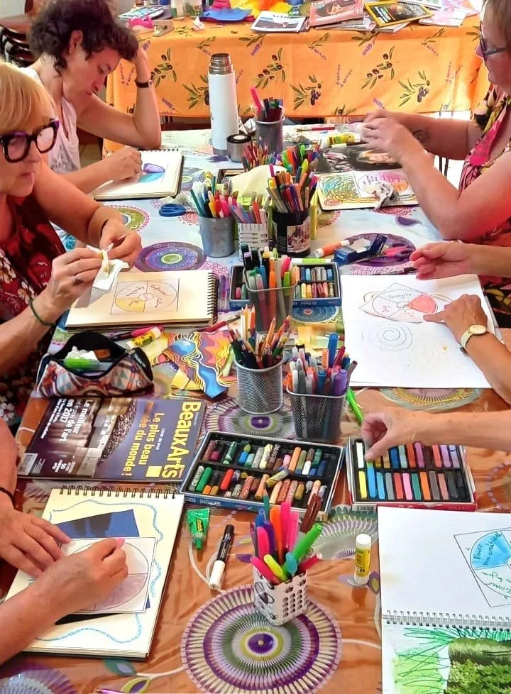 Un groupe de personnes assises autour d'une table en train de faire des activités artistiques avec des crayons, marqueurs et cahiers.