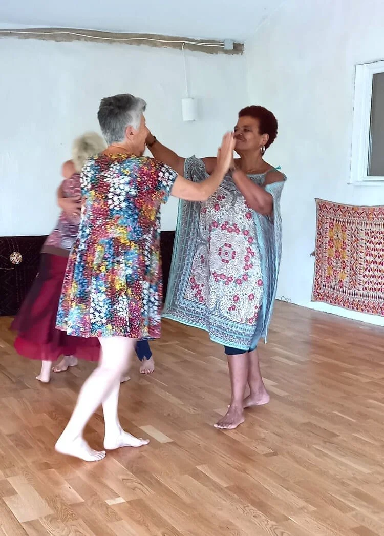 Deux femmes dansent joyeusement dans une pièce avec un parquet en bois, une tapisserie colorée accrochée au mur, et une fenêtre. Elles semblent s'amuser.