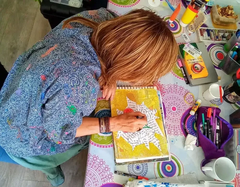 Une femme peint dans un carnet de croquis sur une table colorée, entourée de divers fournitures d'art