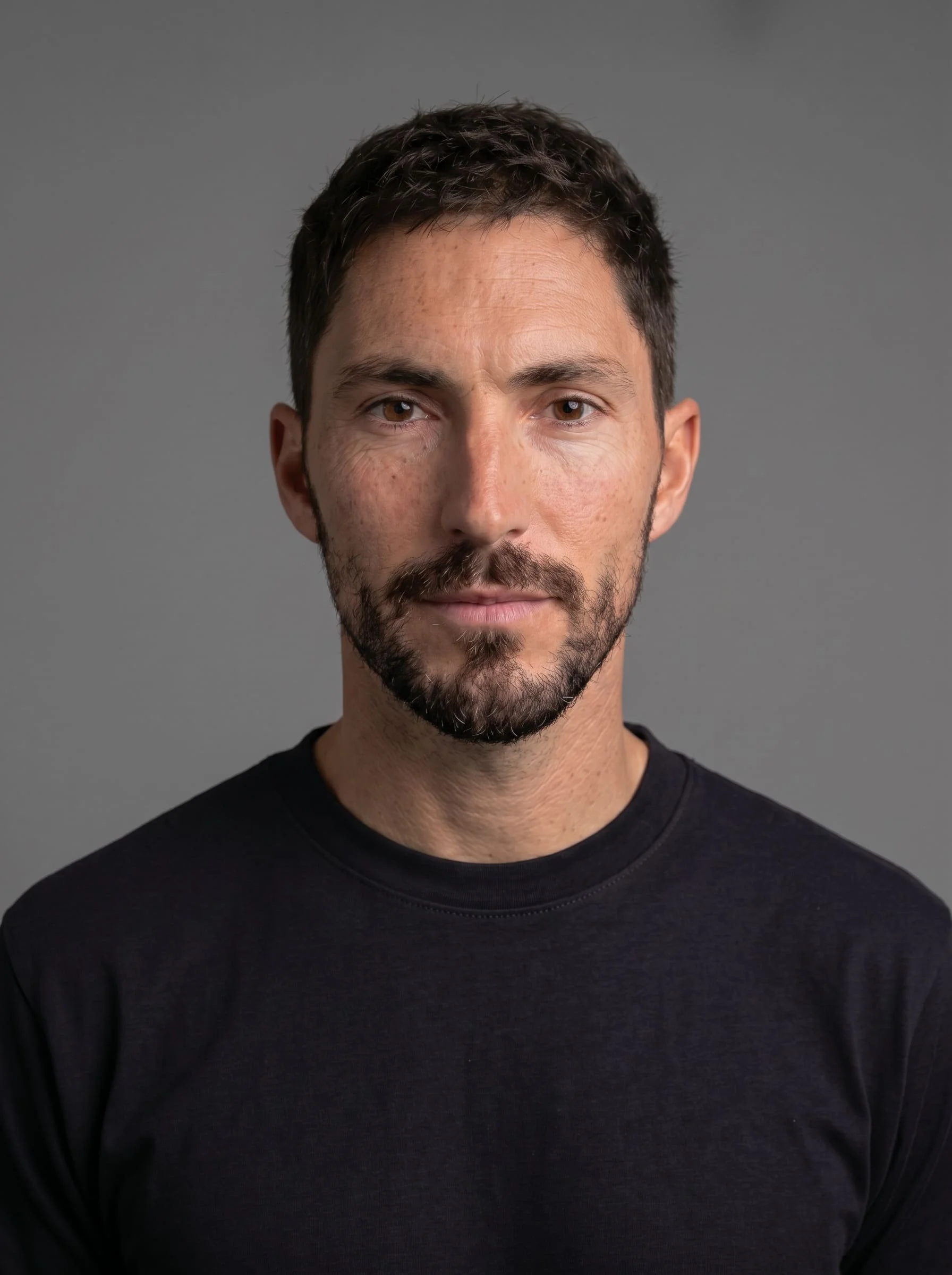 Portrait d'un homme avec barbe brune, cheveux courts, portant un t-shirt noir, contre un fond gris.