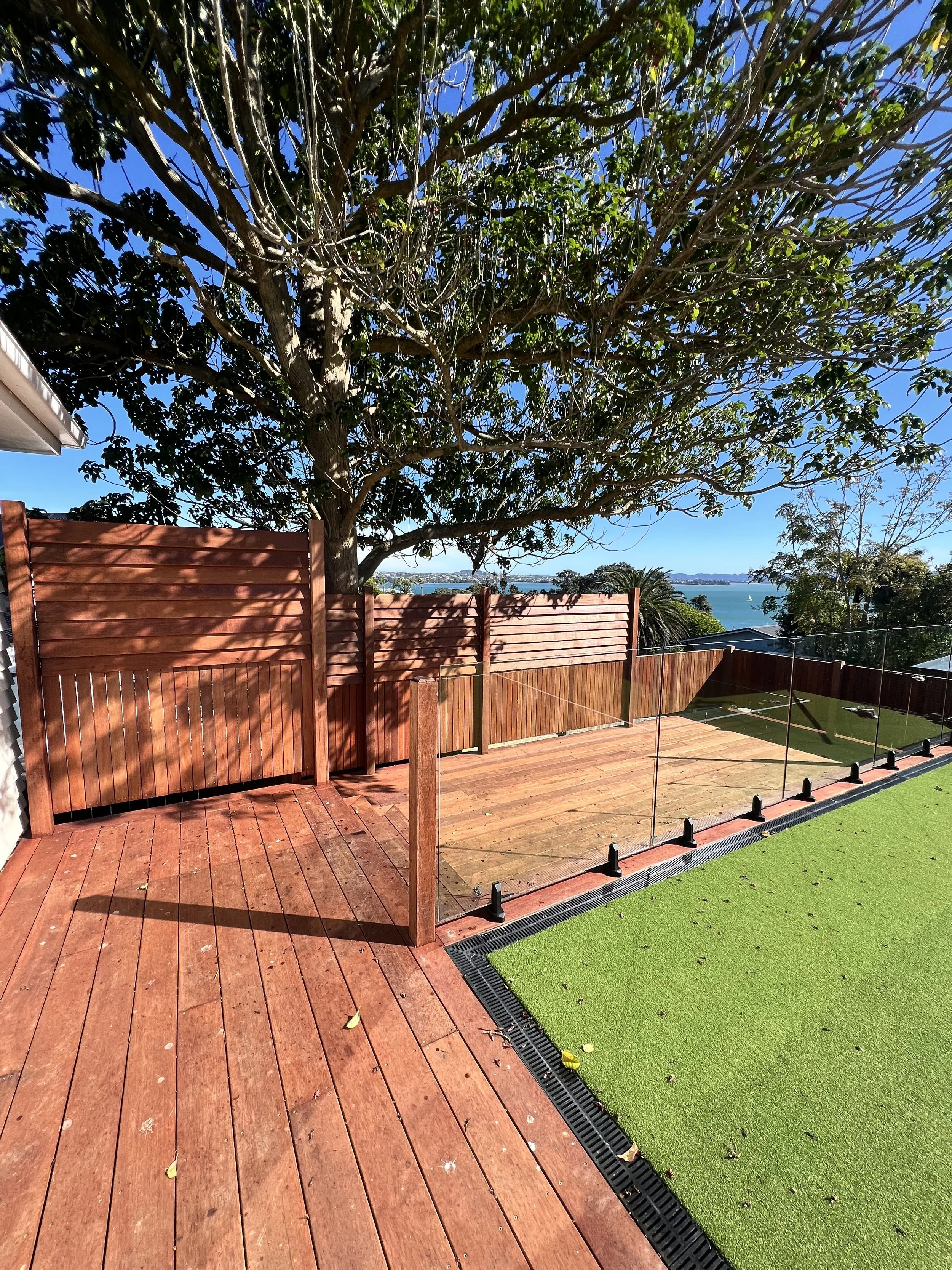 View of a backyard with a wooden deck, a large tree with green leaves, a red fence, a grassy area, and a body of water in the distance under a blue sky.