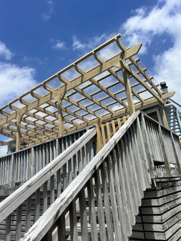 Wooden deck with stairs leading up to a partially built pergola structure under a blue sky with clouds.