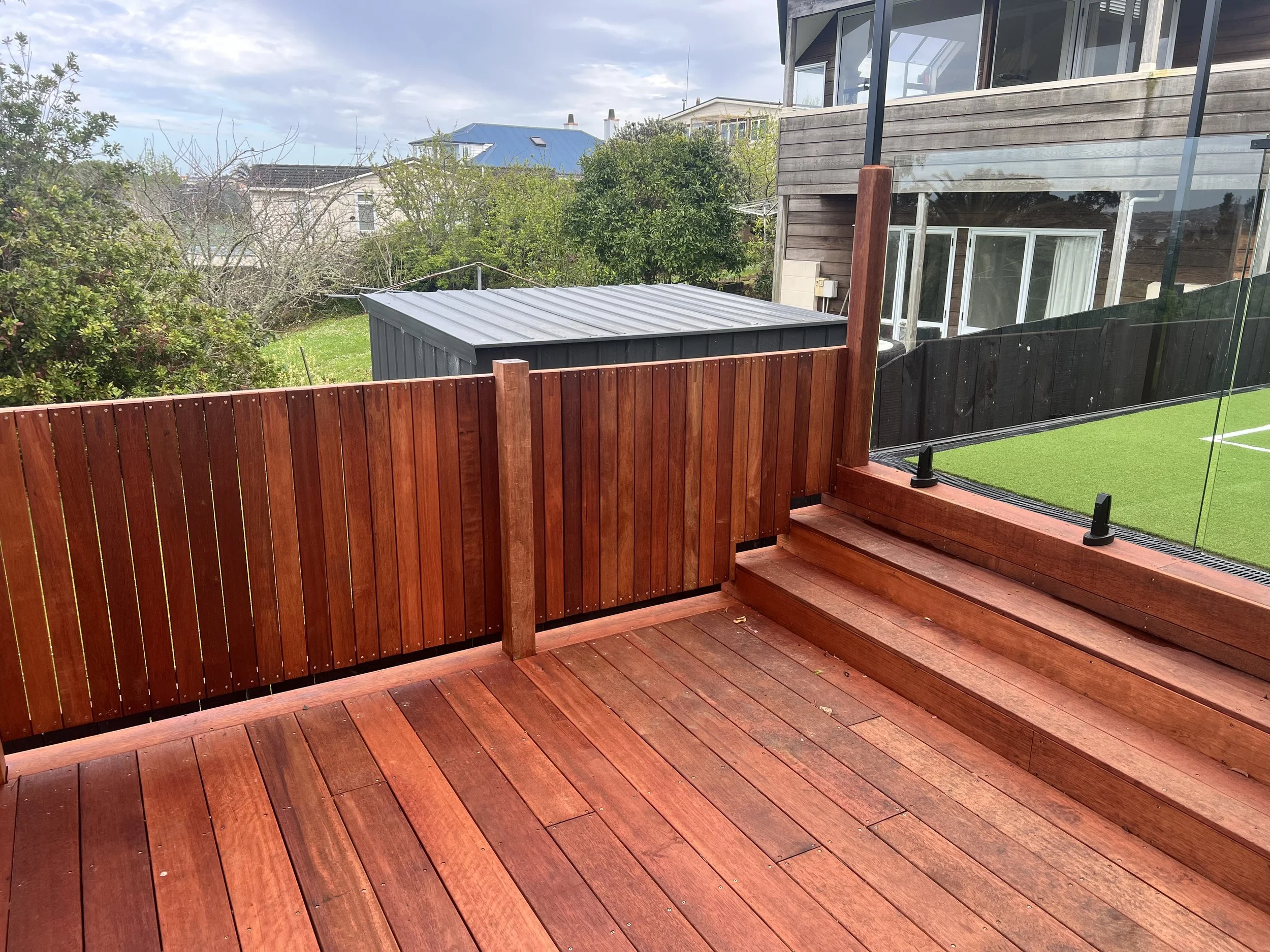 View of a balcony with wooden flooring and a glass railing, overlooking neighboring houses with trees and a cloudy sky.
