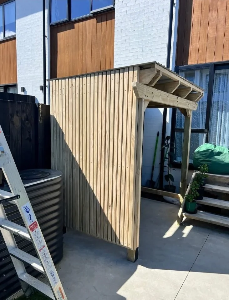 A partially built wooden outdoor structure with vertical slats and a sloped roof, situated on a concrete patio next to stairs with a potted plant, garden tools, and an outdoor air conditioning unit nearby.