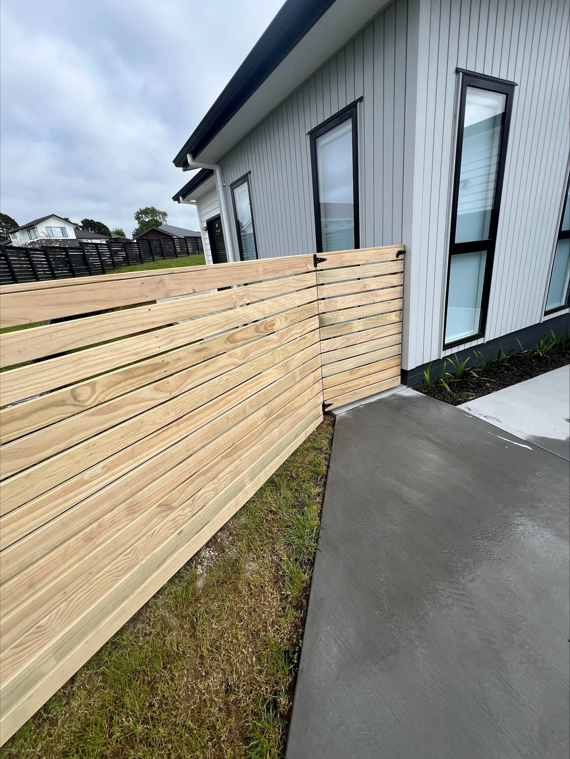 A modern house with vertical siding, large windows, a wooden fence, a concrete walkway, and an overcast sky.