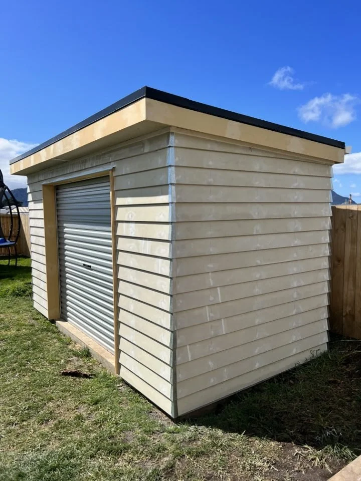 A small beige shed with a roll-up metal door, located in a grassy backyard with a wooden fence and a blue sky.