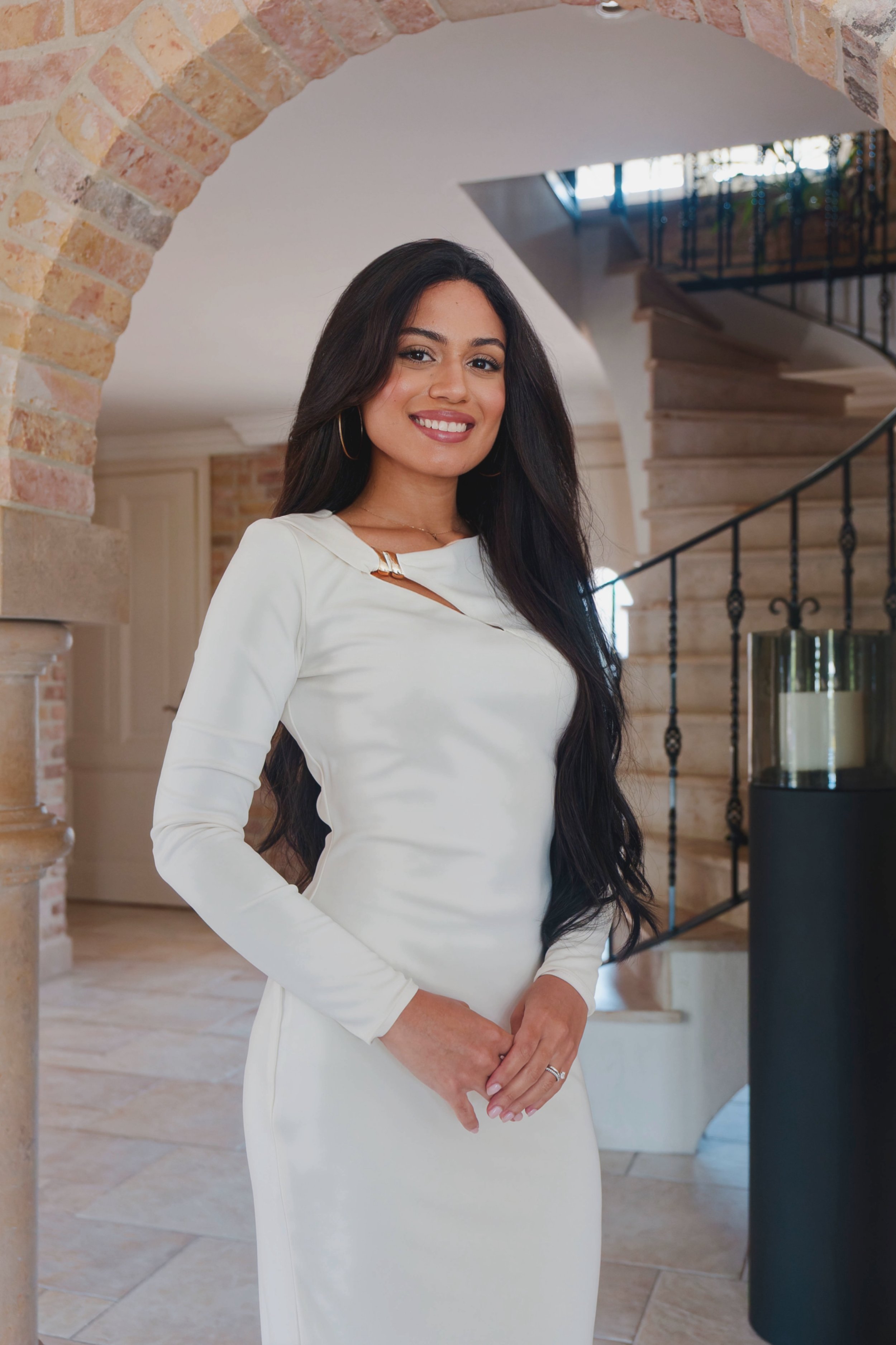 A woman in a white dress standing indoors in front of a staircase with brick archways, smiling at the camera.