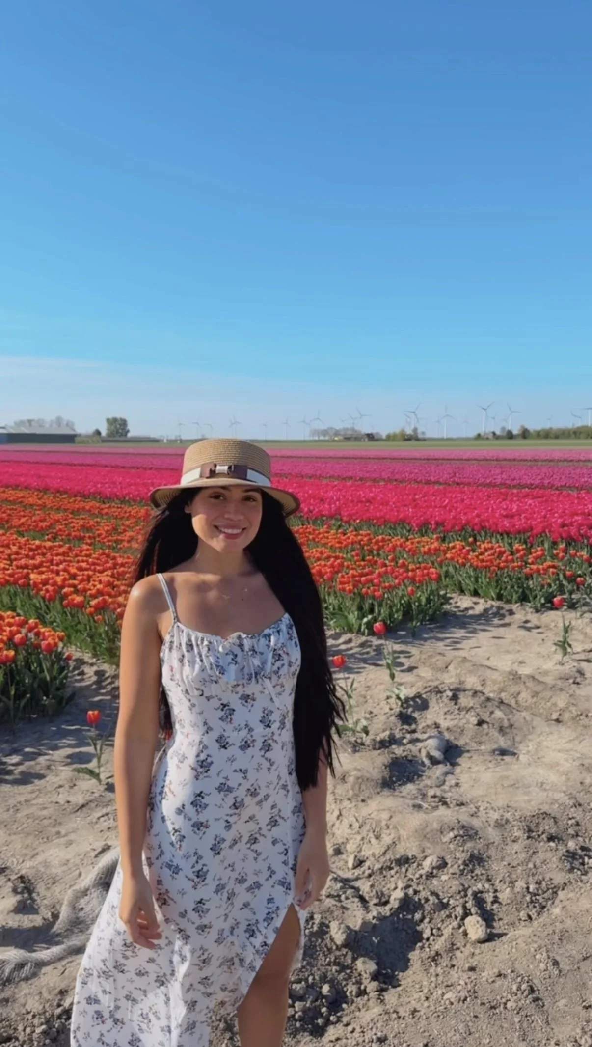 A woman standing in a field of colorful tulips, wearing a white dress with a floral pattern and a wide-brimmed hat, smiling.