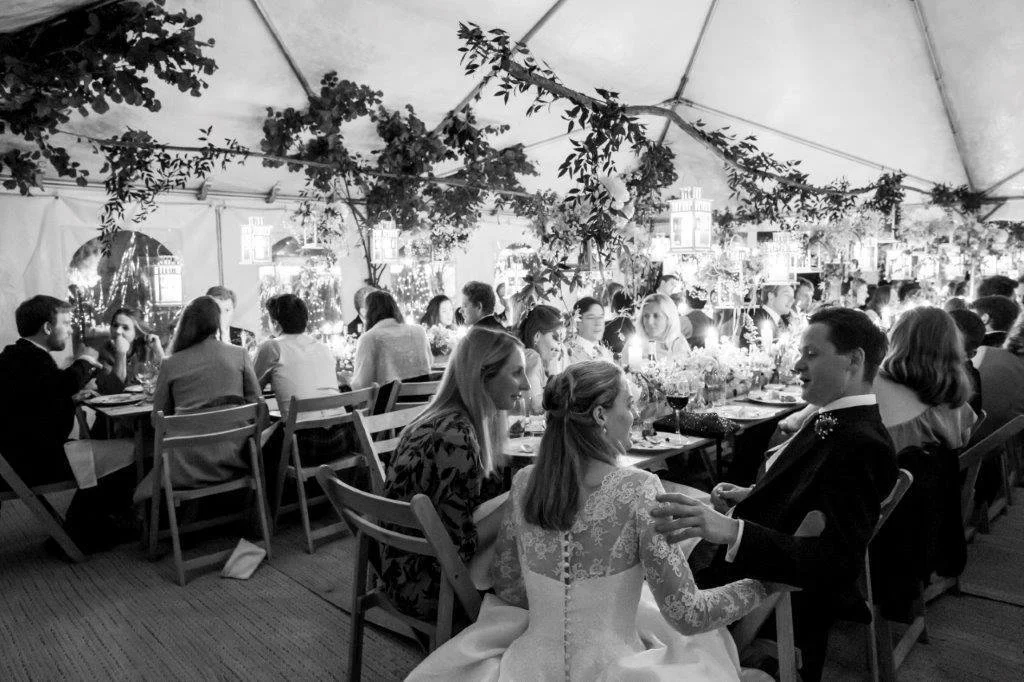 A black and white photo of a wedding reception under a canopy tent decorated with foliage. Guests are seated at long tables enjoying dinner and conversation, with a bride and groom in the center foreground.