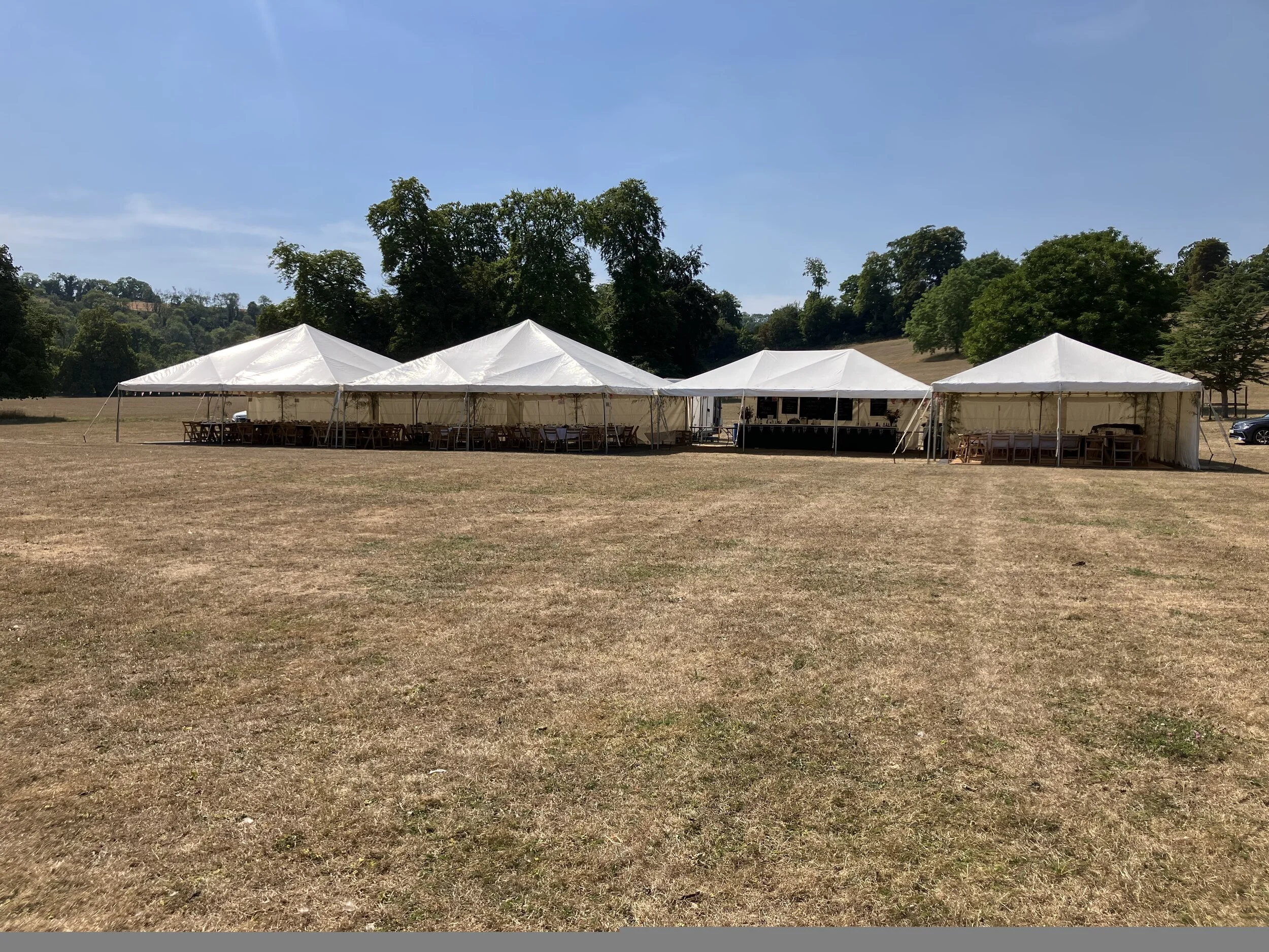 Three white event tents set up on a grassy field with chairs and tables underneath, surrounded by trees and hills under a blue sky.
