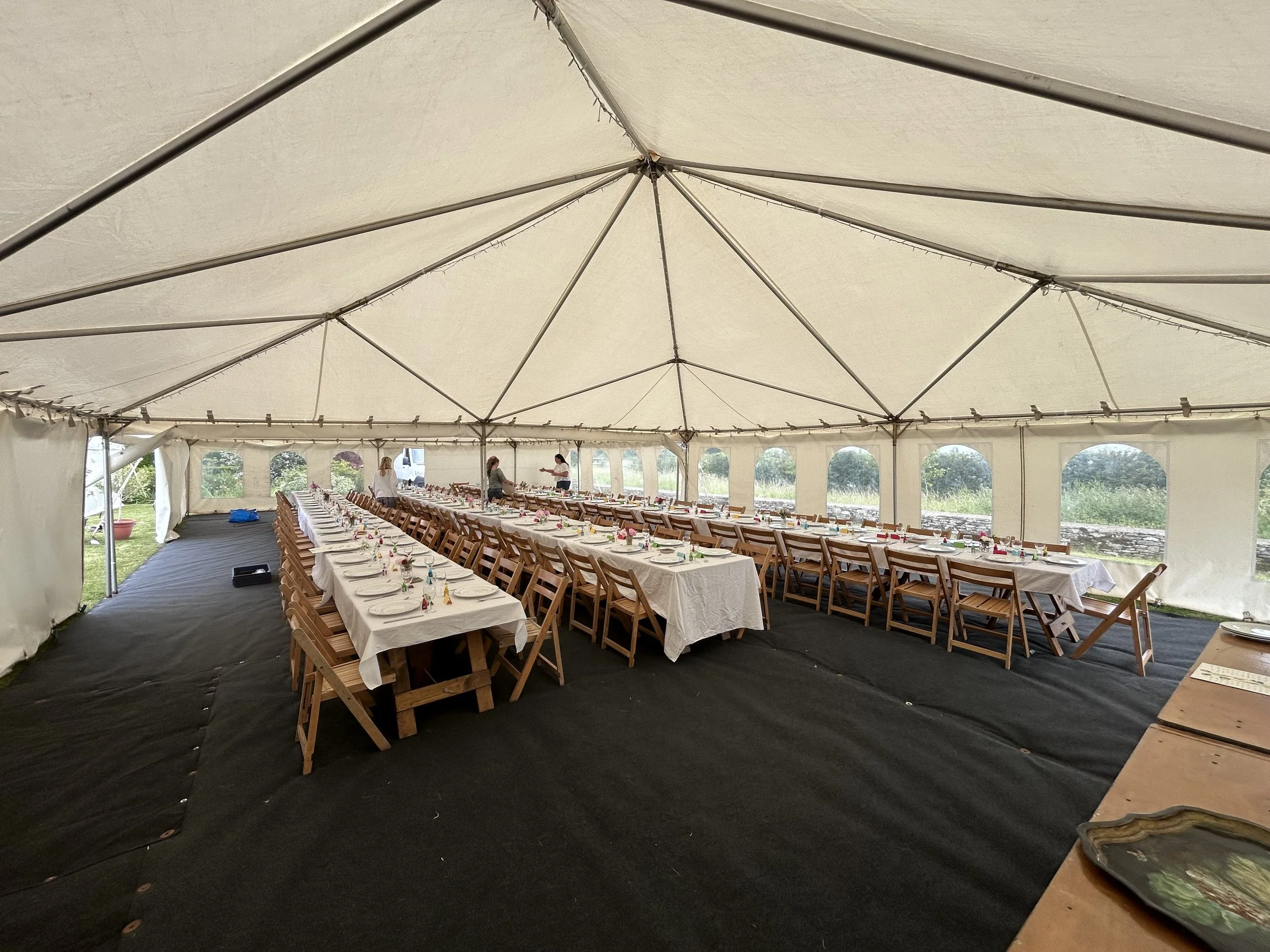 Large white event tent with decorated long banquet tables and chairs inside, set up for a celebration, with some people arranging items and greenery visible outside.