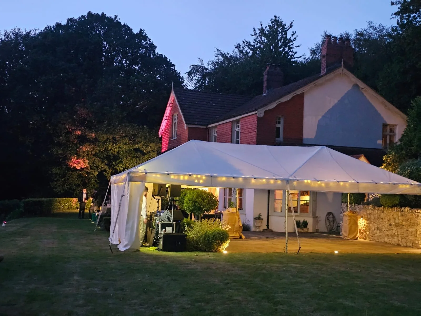 Outdoor evening event setup with a white tent and string lights, set in a yard in front of a large house with a Tudor-style exterior and trees in the background.
