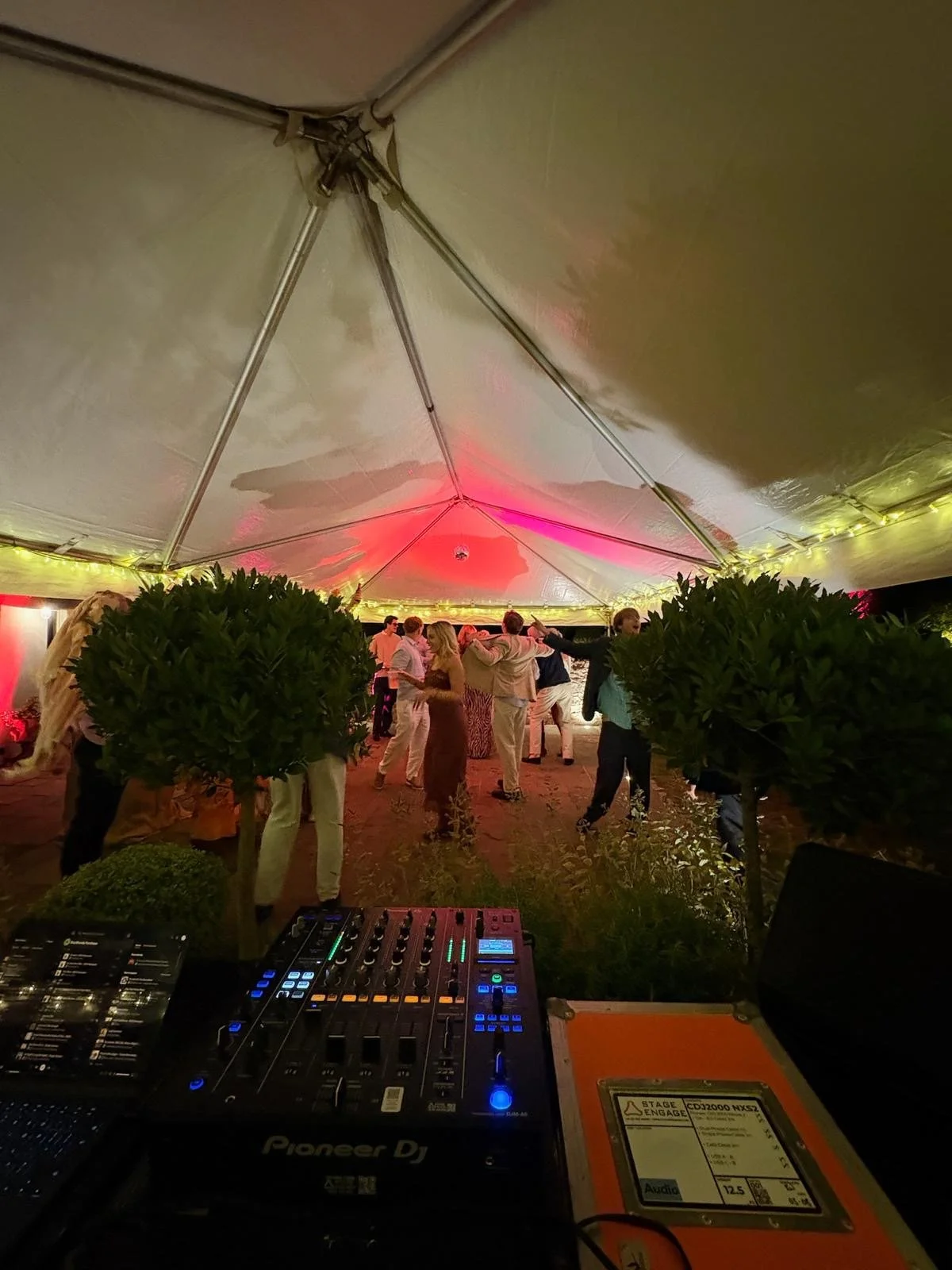 People dancing and socializing under a tent with colorful lighting, with a DJ mixer in the foreground.