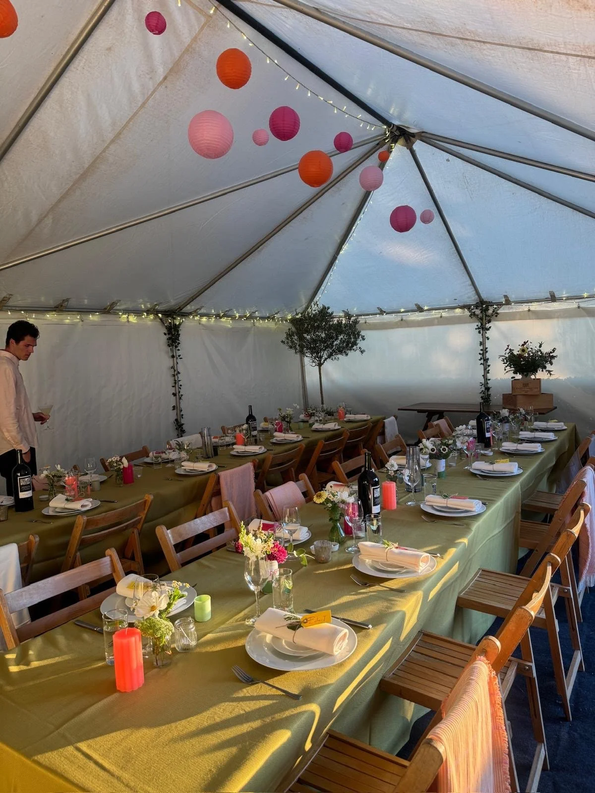 A decorated party or celebration tent with long tables covered in green tablecloths, set with white plates, silverware, glasses, small floral arrangements, candles, and wine bottles. Paper lanterns in pink, orange, and purple hang from the ceiling, w