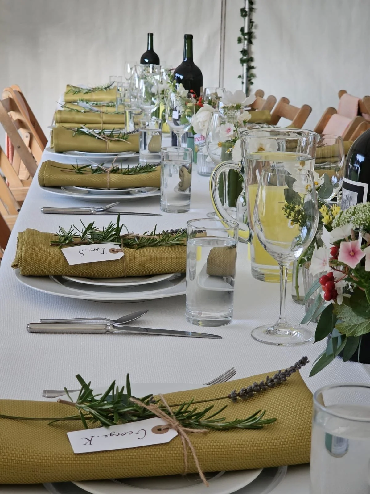 A beautifully arranged dining table with olive-colored napkins rolled and tied with twine and decorated with sprigs of rosemary, set with clear glassware including wine glasses and pitchers, and complemented by small floral centerpieces, white plates