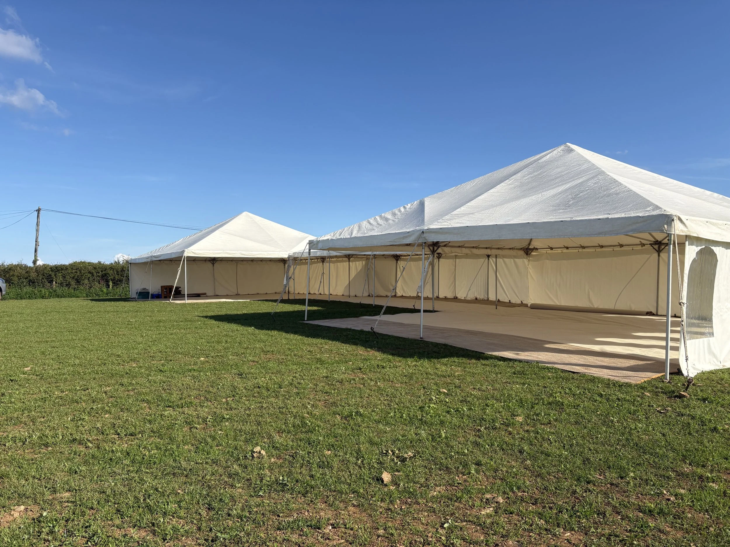 Two large white event tents set up on a grassy field under a bright blue sky with a few clouds. One tent is fully open on one side and the other is partially enclosed. A wooden utility pole with power lines is visible in the background.