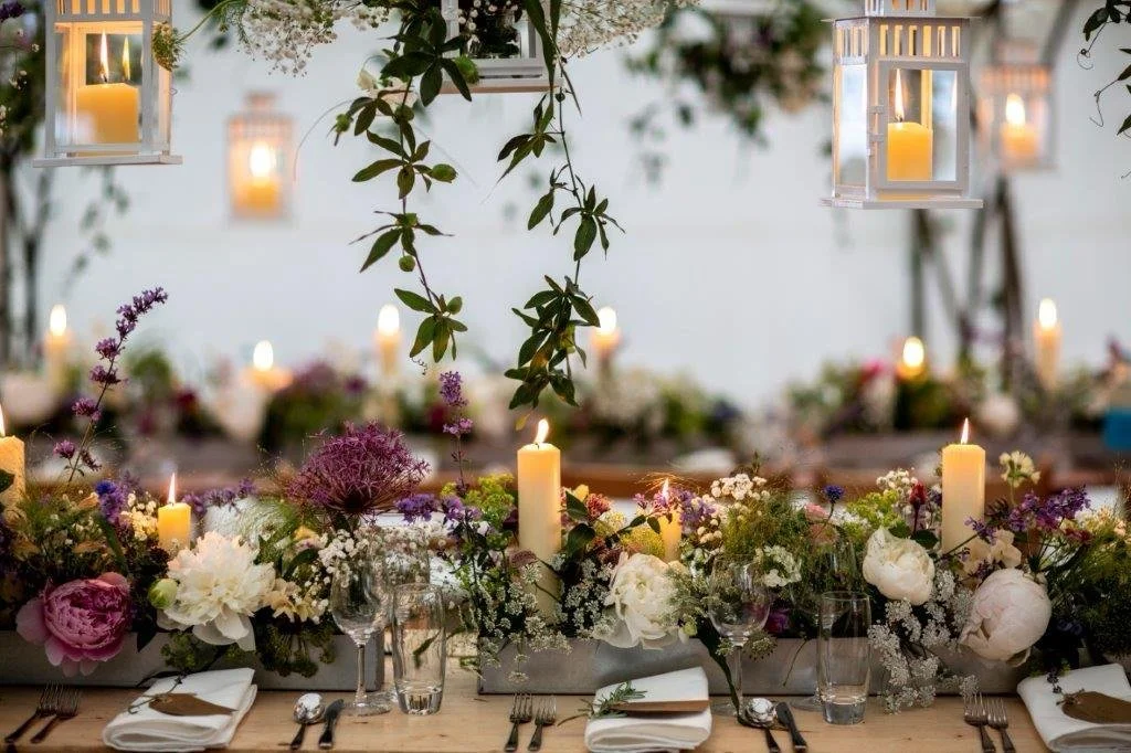 A beautifully decorated outdoor table with a floral centerpiece, surrounded by candles and lanterns, set for a celebration or dinner.