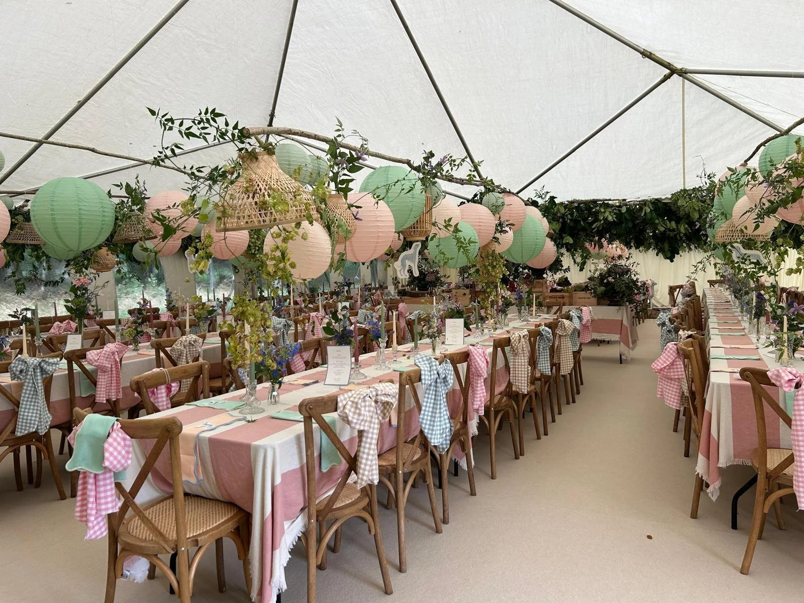 Long dining tables with pink and cream checkered and plain tablecloths decorated with floral centerpieces, surrounded by wooden chairs with pink and blue checkered bows, under hanging pastel paper lanterns and greenery in a tented setting for a celeb