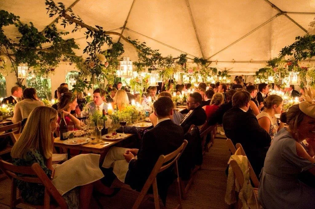 People dining at an elegant event tent decorated with greenery and hanging lanterns, with warm lighting and tables set with wine bottles and glasses.