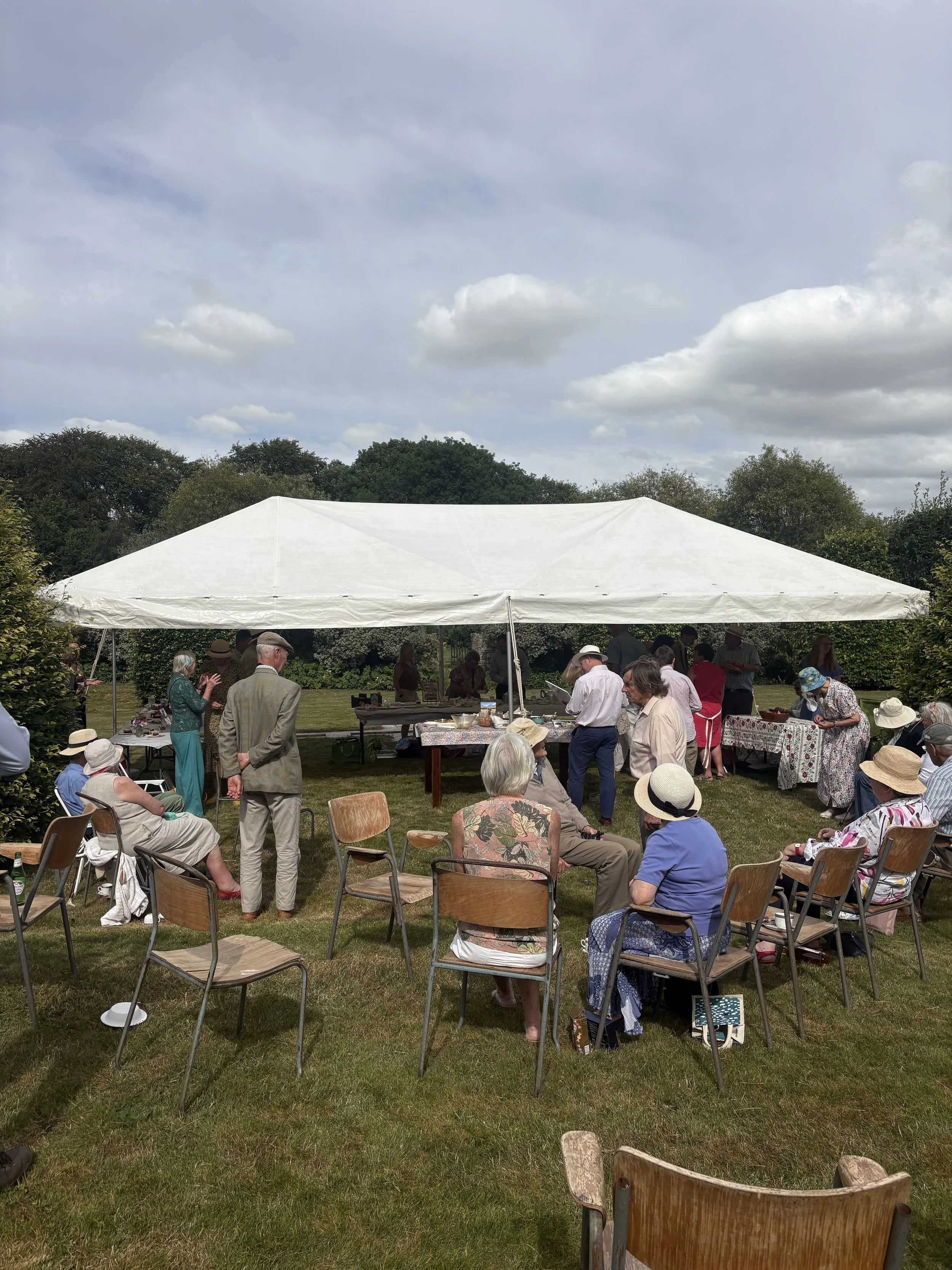 People gathered outdoors under a white canopy, sitting and standing on grass, with trees and cloudy sky in the background.