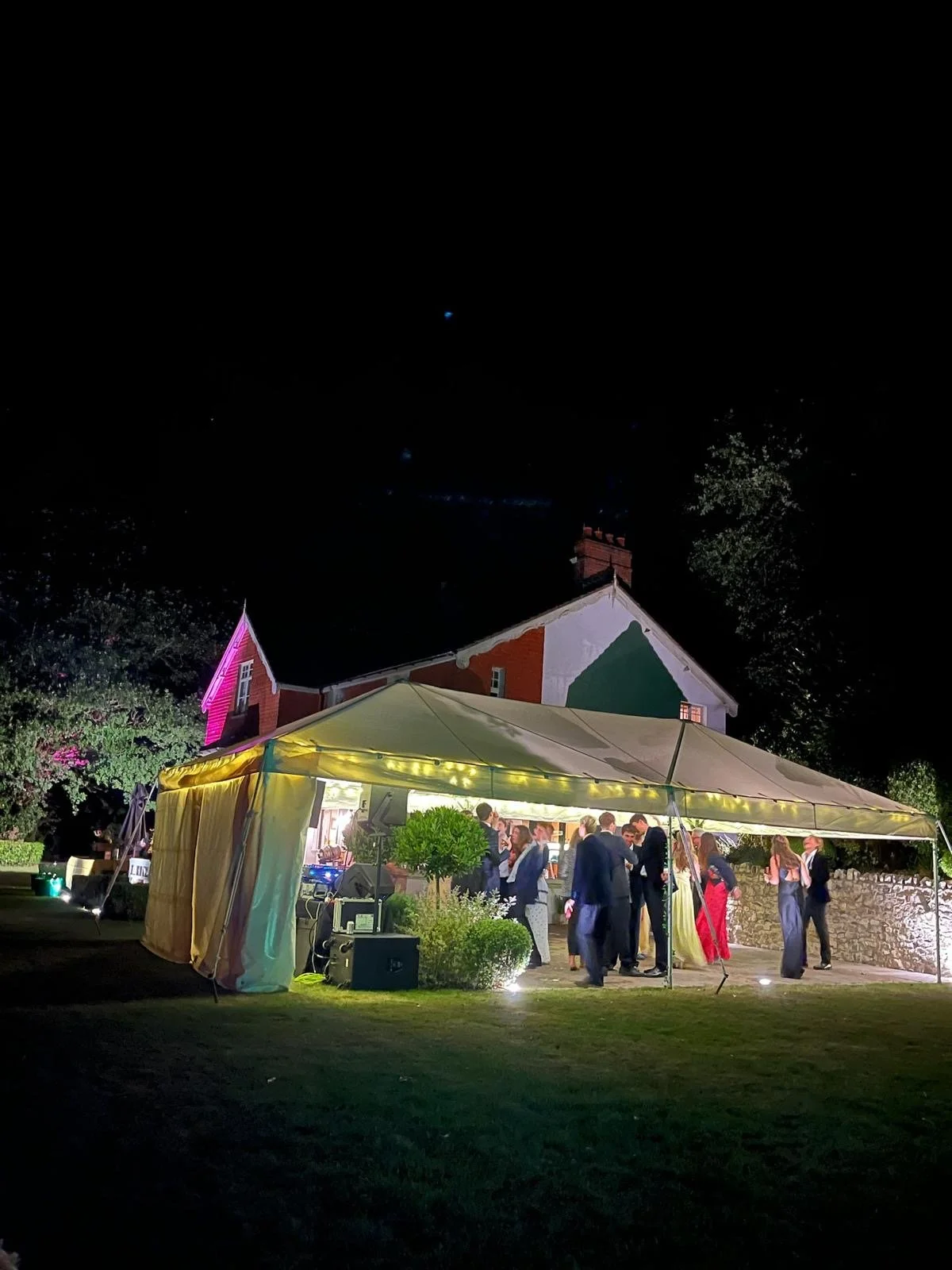 People dancing and socializing under a yellow canopy at an outdoor evening event, with a barn in the background illuminated by colorful lights.