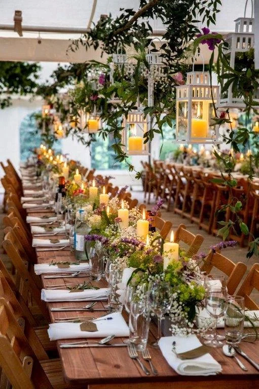 Long banquet table decorated with flowers, candles, and lanterns, set for a formal event in a tented outdoor space.