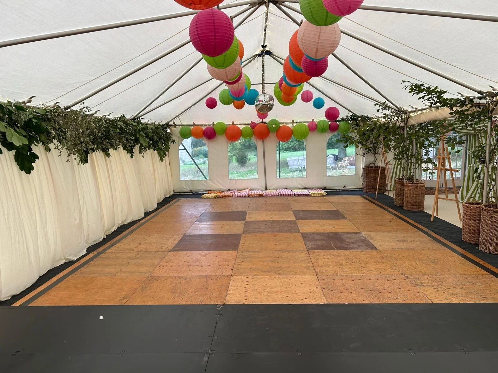 Empty decorated event tent with multicolored paper lanterns hanging from the ceiling, green leafy garlands along the sides, and a wooden dance floor, with outdoor greenery visible through the windows.