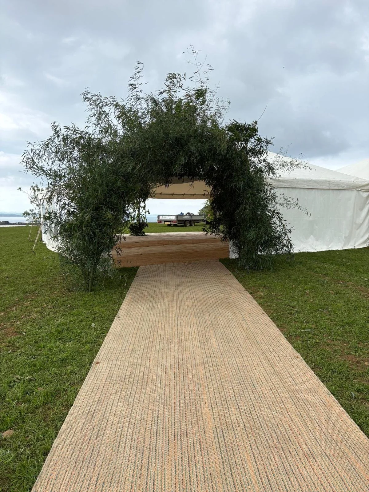 Outdoor event space with a beige pathway leading to a stage framed by lush greenery, under cloudy sky, with a tent on the right and a trailer in the background.