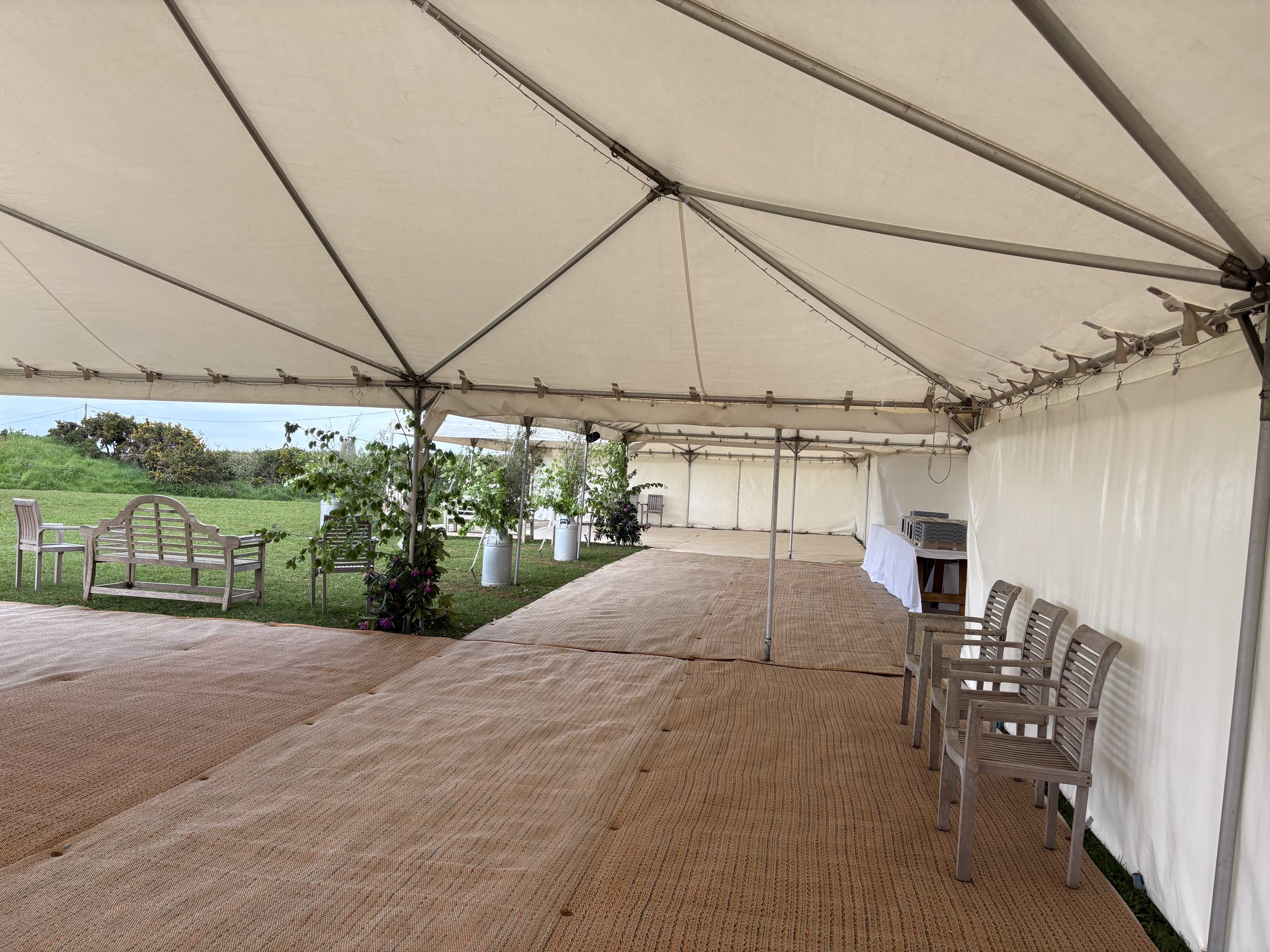 Inside of a large white event tent with chairs along the side, a beige ground covering, and some outdoor landscape visible through the open side of the tent.