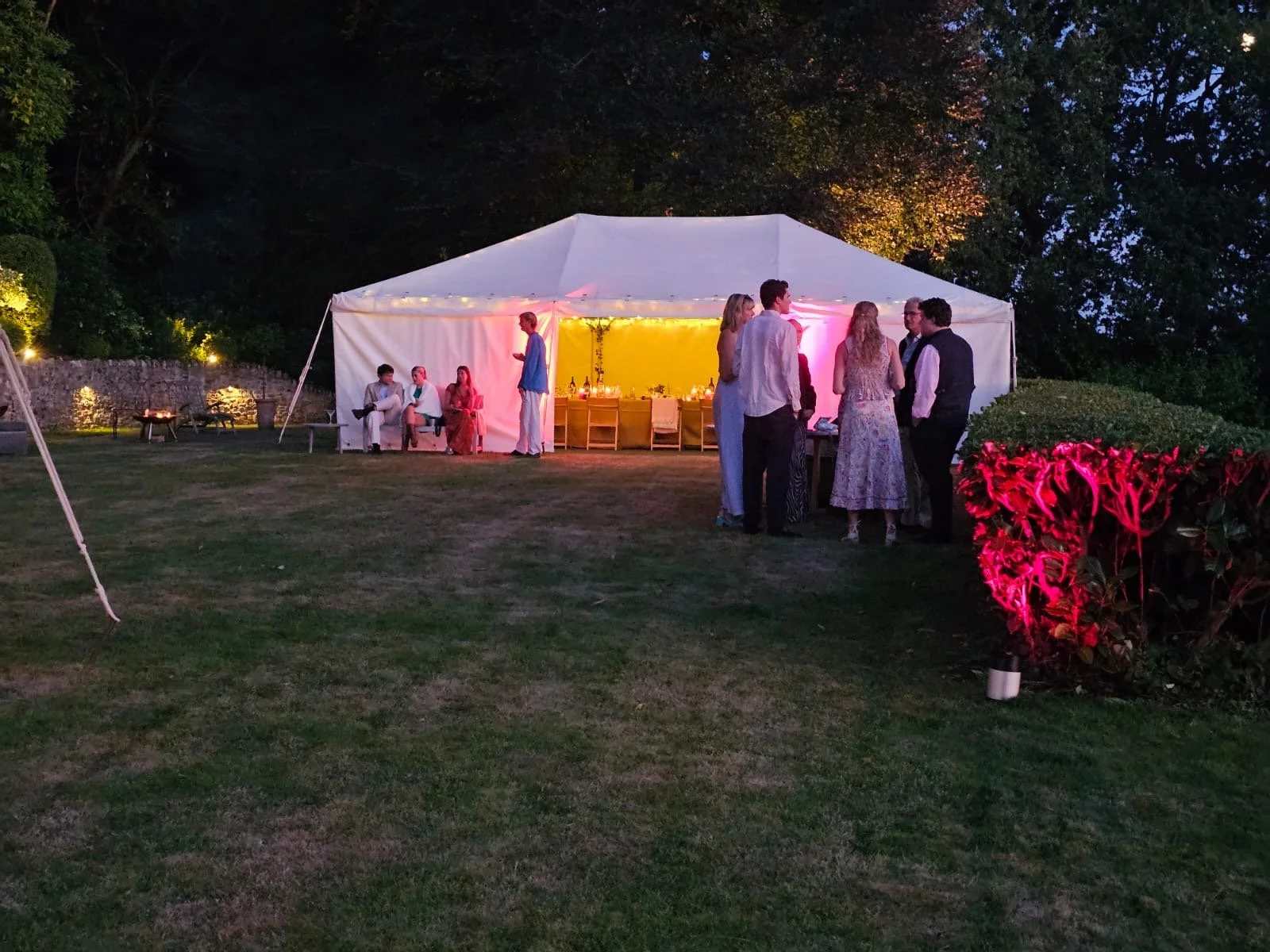 Outdoor evening gathering with a white event tent, people socializing and sitting, decorated with string lights and pink lighting, set on a grassy area with trees and bushes.