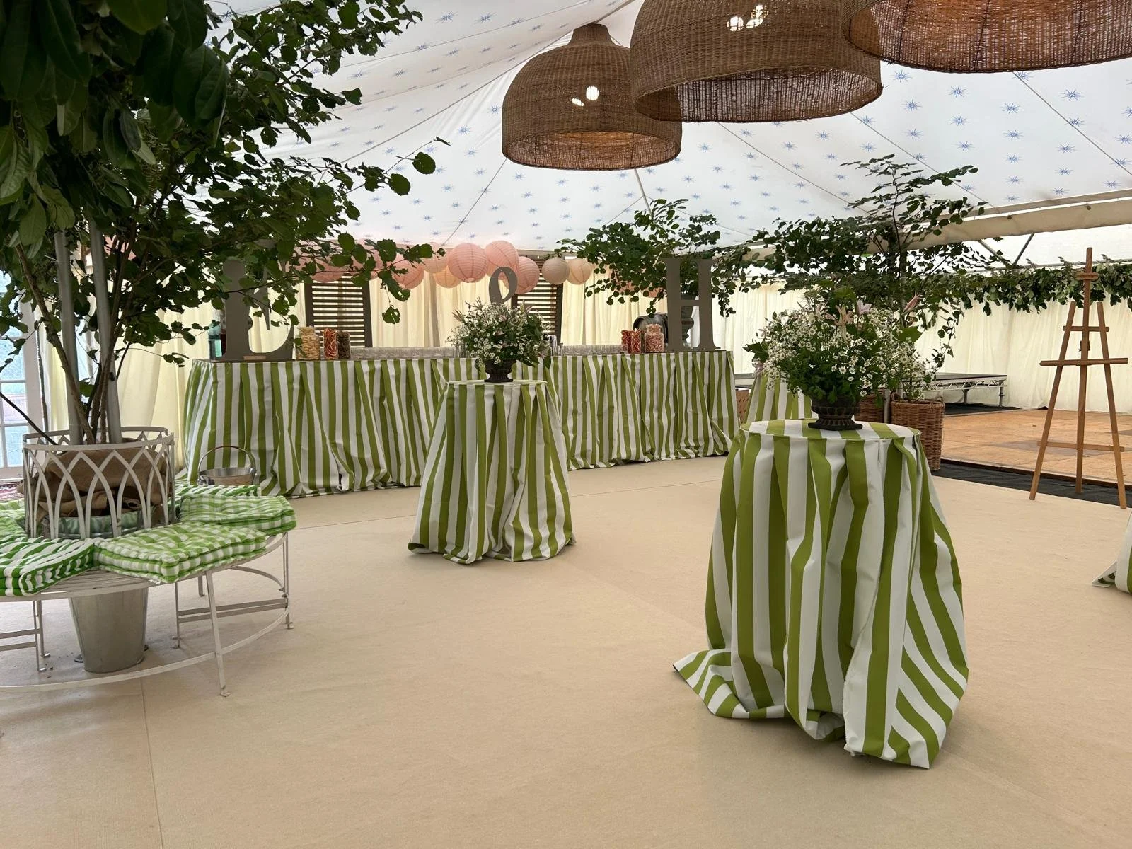 Event space decorated with green and white striped fabric covering tables and drapes, with potted plants and hanging paper lanterns, set up for a celebration or gathering.