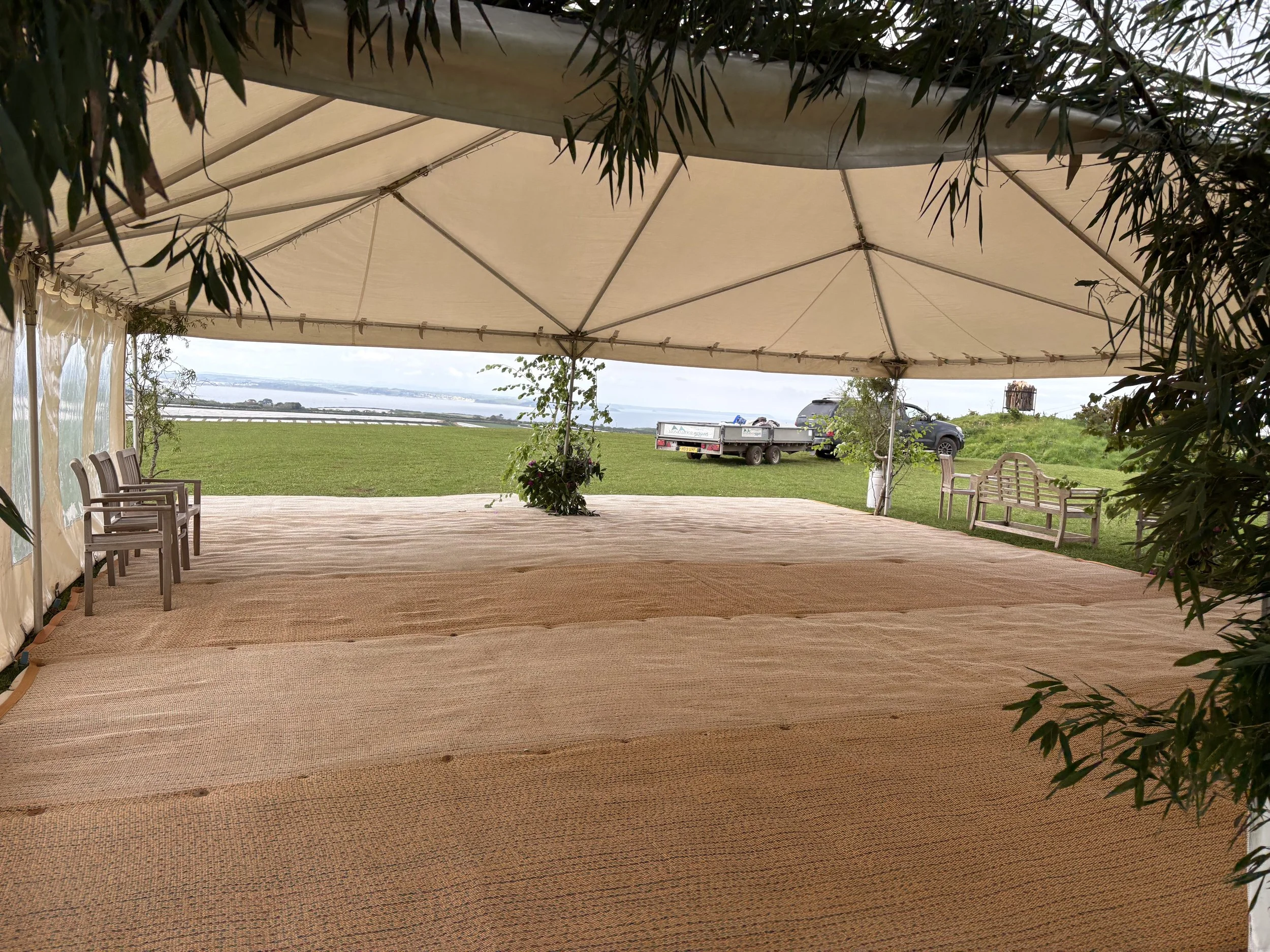 A large outdoor event tent with a beige canopy, set up on a grassy area overlooking a body of water and distant land. There are chairs along the side and a potted plant in the middle, with a truck and vehicle visible in the background.