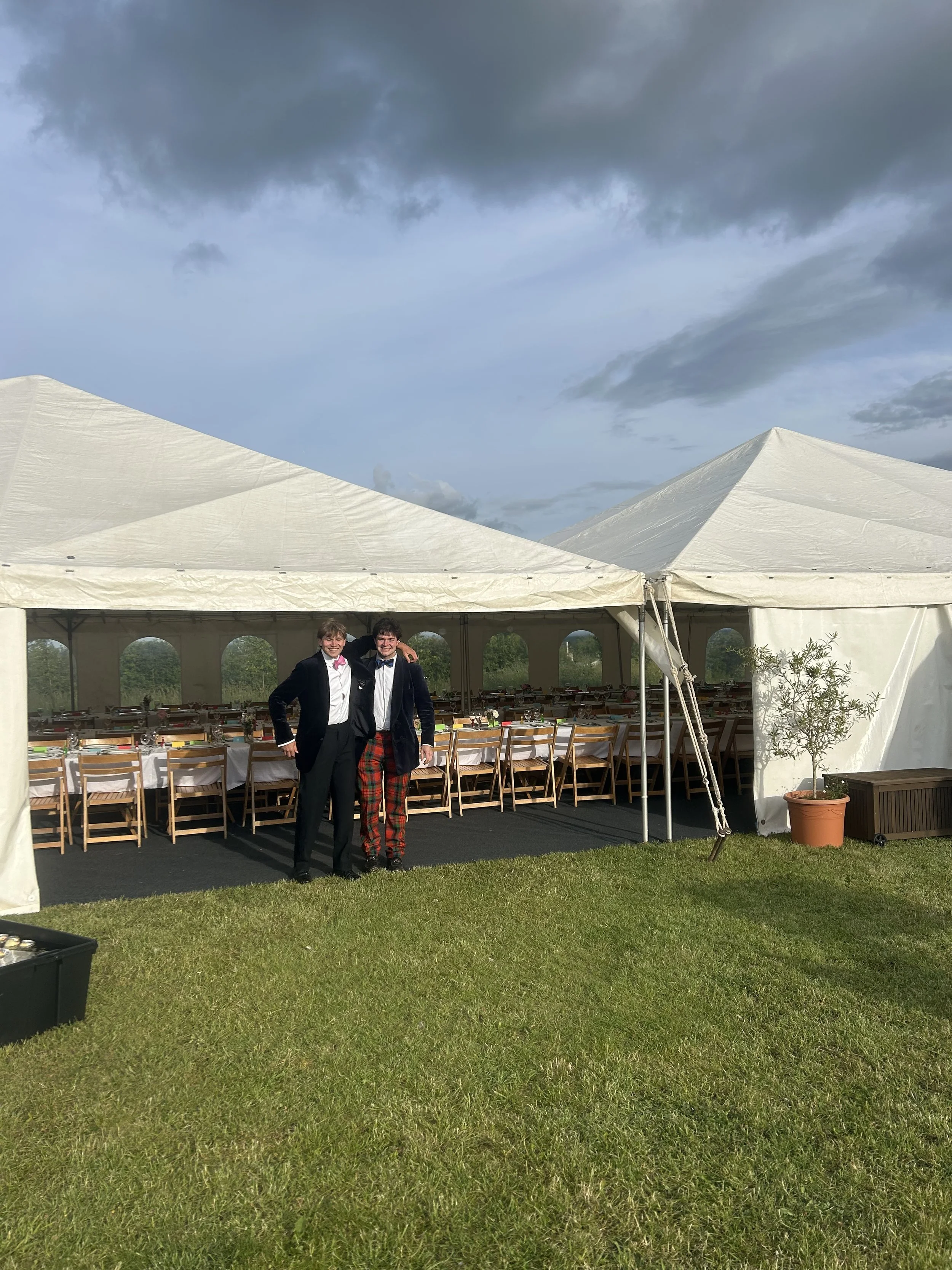 Two men dressed in tuxedos with one in a kilt stand in front of a large white event tent on a grassy area under a cloudy sky. The tent is set up for a celebration or gathering, with tables and chairs inside.