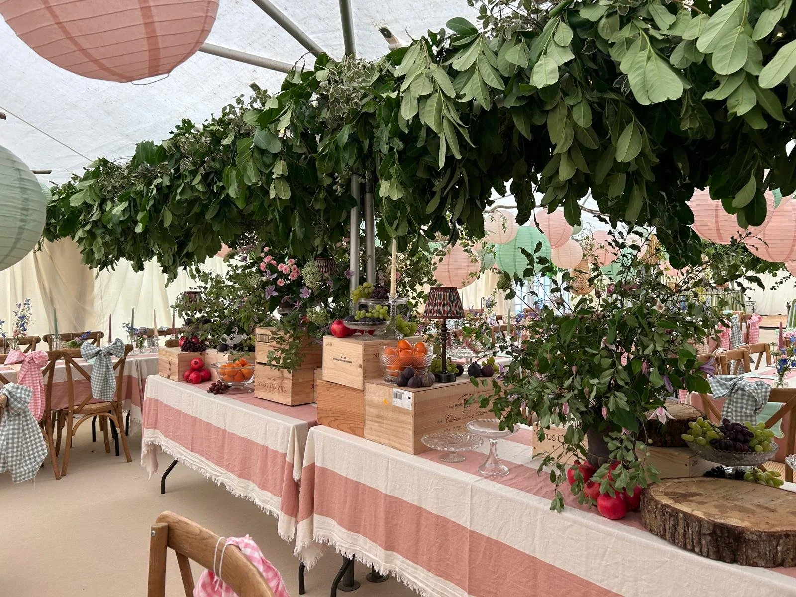 Table decorated with potted greenery, fruit bowls, and wooden crates, in a tented setting for a celebration, with pink, green, and white paper lanterns hanging overhead.