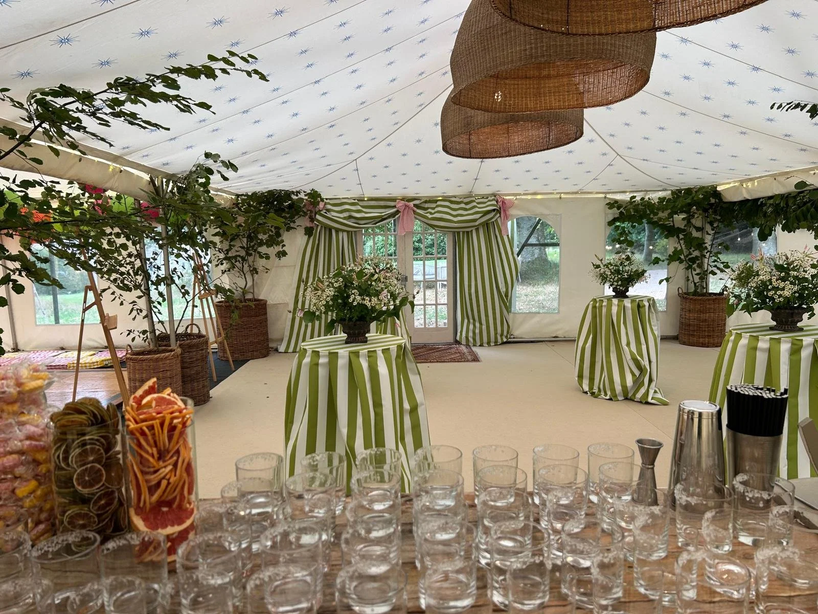 A decorated event tent with striped green and white drapes and tablecloths, plants, and flowers. The ceiling is adorned with hanging woven lampshades and string lights. On the table in the foreground, there are glasses, a tray of candies, and drinkwa
