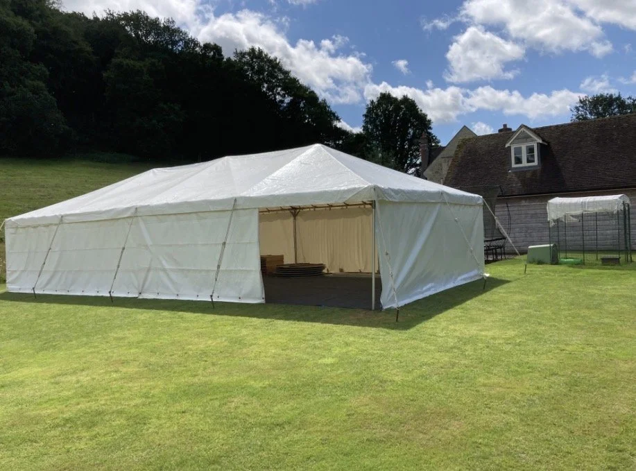 A large white outdoor event tent set up on a grassy lawn, with an open side revealing stacked chairs inside, neighboring a house and a small greenhouse.